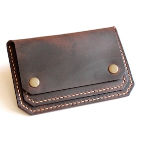 Peut inclure: Un porte-cartes en cuir marron foncé doté d'un rabat rectangulaire fixé par deux boutons-pression en métal couleur laiton. Les bords sont renforcés par des coutures apparentes de couleur claire, donnant un aspect structuré à cet accessoire.