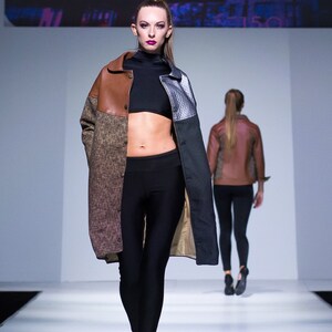 May include: A model walks the runway wearing a brown, black, and silver patterned jacket with a black top and black leggings. The model is wearing black heels.