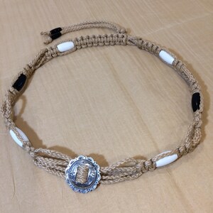 May include: A tan rope belt with white and black beads and a silver concho. The concho has a floral design.