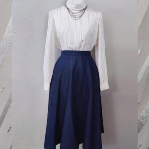 Könnte beinhalten: Eine weiße Langarmbluse mit vertikalen Streifen und hohem Kragen, kombiniert mit einem dunkelblauen Jeansrock. Eine mehrreihige silberne Halskette schmückt den Ausschnitt. Das Outfit wird auf einer Schaufensterpuppe präsentiert.