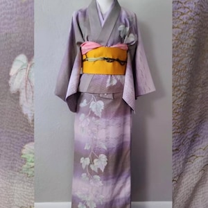 May include: A lavender kimono with a grey collar and sleeves, displayed on a mannequin. The kimono features a yellow obi with a pink sash and a vine leaf pattern. The fabric has a subtle wave texture.