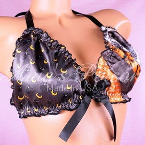 Puede incluir: Bralette de satén negro con estampado de gato y luna creciente negro y amarillo. La bralette tiene un lazo de cinta negro en el centro y una tira de halter negra.