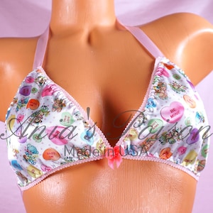 May include: A white satin triangle bra with pink straps and a pink bow in the center. The bra is covered in a pattern of candy hearts with various messages, including "Be Mine", "True Love", and "Love My", on a white background with pink polka dots.