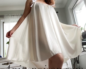 Könnte beinhalten: Ein weißes Satin-Schlafkleid mit dünnen Trägern und einem weißen Schleifendetail am Ausschnitt. Das Kleid ist fließend und hat eine lockere Passform.