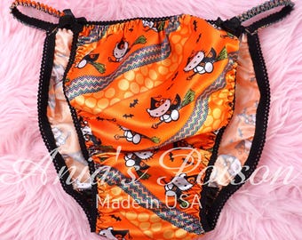 May include: Orange satin underwear with a Halloween-themed print featuring a cartoon cat riding a broom, bats, and polka dots. The edges are trimmed with black lace and small black bows. The text "Made in USA" is visible.