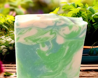 May include: A rectangular bar of handmade soap with a marbled design. The soap features swirls of green, blue, and white, creating a natural, organic appearance. The soap is set against a blurred background of greenery and sunlight.