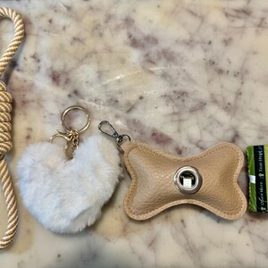 Könnte beinhalten: Eine Sammlung von Haustierzubehör, darunter eine beige-weiße Seilleine, ein flauschiger, herzförmiger Schlüsselanhänger, ein beigefarbener Knochen-Beutelspender aus Leder und eine Rolle grüne Müllbeutel.