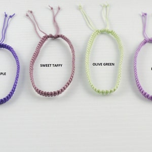May include: Six different colors of braided friendship bracelets. The colors are green, purple, pink, olive green, lilac, and lime green.