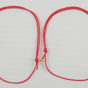May include: Two red string bracelets with gold beads. The bracelets are tied in a knot at the end.