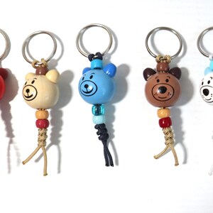 May include: A collection of seven bear-shaped keychains in various colors, including burgundy, red, tan, blue, brown, white, and pink. Each keychain features a metal ring, a wooden bear head, and a beaded tassel. The bears have painted facial features.