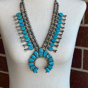 Puede incluir: Un collar nativo americano de turquesa y plata con un gran colgante en forma de media luna. El collar está hecho de múltiples hileras de cuentas de plata y presenta piedras turquesa de diferentes tamaños.