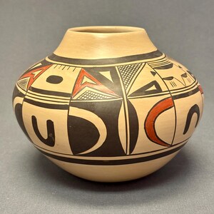 May include: A beige ceramic pot with a rounded body and a narrow neck. The pot is decorated with black, red, and white geometric patterns, including triangles, curves, and lines. The design is symmetrical and covers the majority of the pot's surface.