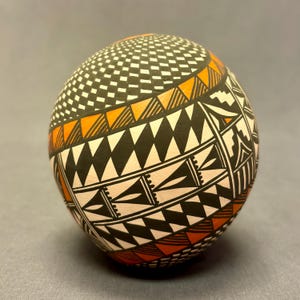 May include: A spherical, hand-painted ceramic vessel with intricate geometric patterns. The design features black, white, and orange triangles, diamonds, and other shapes. The top section has a repeating grid pattern. The vessel is a decorative art piece.