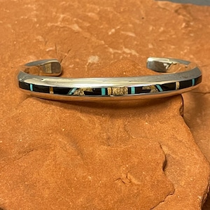 Op de afbeelding: Een zilveren manchetarmband met een zwart, goud en turkoois inlegontwerp. De armband heeft een rechthoekige vorm met afgeronde randen en wordt weergegeven op een roodbruin oppervlak.