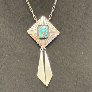 May include: A silver-toned necklace with a turquoise stone set in a decorative pendant. The pendant has a sunburst design and two dangling geometric shapes. The chain is a simple link style.