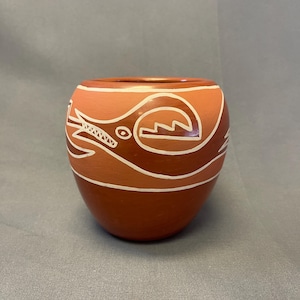 May include: A hand-painted, round ceramic pot with a reddish-brown base and a lighter orange top. The pot features a white, stylized design of a creature and geometric shapes. The design wraps around the pot.