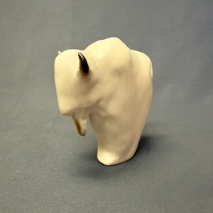 May include: A cream-colored ceramic bison sculpture with black-tipped horns and a tan-colored beard. The bison is standing on a gray surface. The sculpture is a decorative item.