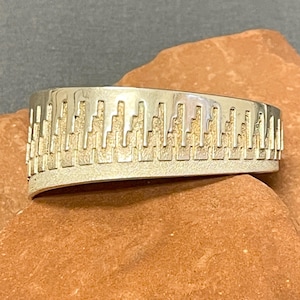 May include: A silver-toned ring with a cityscape design. The ring has a wide band with a textured surface and a series of vertical rectangular cutouts along the top edge, creating a skyline effect. The ring is displayed on a brown surface.