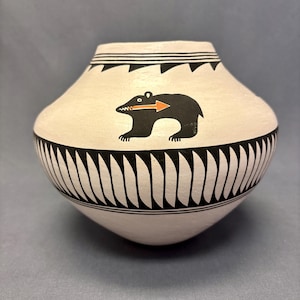 May include: A cream-colored ceramic pot with black geometric designs and a black bear illustration with an orange arrow. The pot has a rounded body and a slightly flared rim. The bear illustration is centered on the pot.