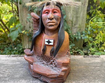 May include: A detailed bust of a Native American figure, featuring a headdress with a feather and straw-like accents. The sculpture has a reddish-brown complexion, with painted facial details and long black hair. A decorative emblem is on the chest.