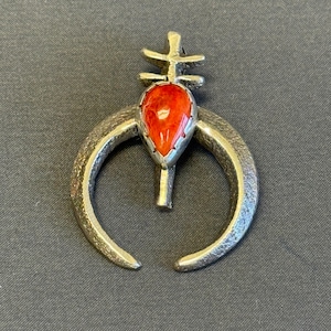 May include: A silver-toned pendant with a teardrop-shaped, vibrant red stone at its center. The pendant features a crescent-shaped design with a cross-like element at the top. The metal has a textured appearance, and the overall design is intricate.