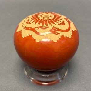 May include: A red pottery bowl with a carved design of a bird and a sunburst. The bowl is on a clear glass stand.