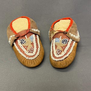 May include: A pair of brown suede moccasins featuring red fabric trim around the openings and intricate beadwork on the vamps. The beadwork displays a floral-inspired pattern in white, blue, and red glass beads against a reddish-brown background.