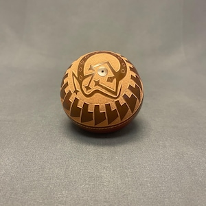 May include: A round, hand-carved wooden object with a light brown central design and a darker brown geometric pattern. The object has a small, silver-colored metal piece in the center of the light brown design.
