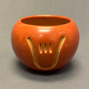 May include: A small, round, terracotta-colored ceramic bowl. The bowl features a carved design on the front, resembling a stylized face or symbol. The interior of the bowl is a darker shade of brown. The bowl is likely handmade.