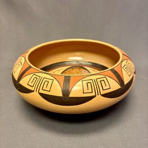 May include: A hand-painted ceramic bowl with a wide, shallow design. The bowl features geometric patterns in black, orange, and tan. The interior has a textured, speckled design. The bowl is approximately 3 inches tall and 8 inches in diameter.