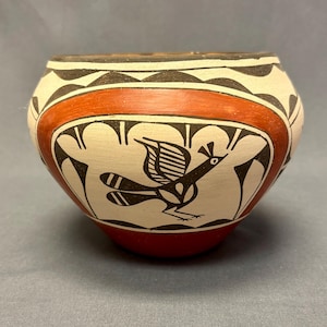 May include: A small, white ceramic pot with a red band and black geometric designs. A black and white bird is painted on the front of the pot.