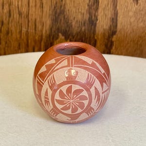 May include: A small, round, terracotta-colored pottery vessel with a carved, cream-colored design. The design includes geometric shapes, a turtle, and a central floral motif. The vessel has a small opening at the top and sits on a white surface.