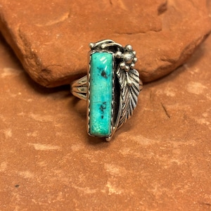 May include: A silver ring featuring a rectangular turquoise stone. The ring has a decorative floral and feather design on the side. The turquoise stone has dark blue and black inclusions. The ring is set on a reddish-brown surface.