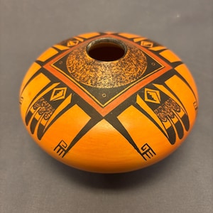 May include: A round, orange ceramic vessel with a small opening at the top. The vessel is decorated with black geometric patterns, red accents, and a textured, speckled design around the opening. The design is symmetrical and appears to be hand-painted.