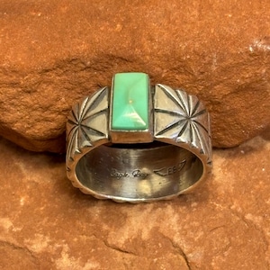 May include: A silver ring with a rectangular, light green stone. The ring has a wide band with starburst designs on the sides. The words "Eagle Boy" are engraved on the inside of the band.