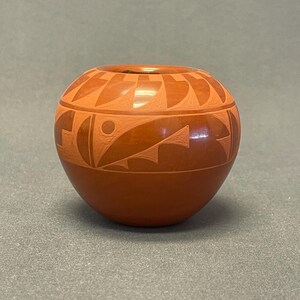 May include: A round, terracotta-colored ceramic pot with a small opening at the top. The pot features geometric designs carved into the surface, including triangles, lines, and curved shapes. The design is lighter in color than the base.