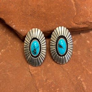 May include: A pair of oval-shaped stud earrings. Each earring features a vibrant turquoise stone set in a silver-colored metal frame. The frame has a sunburst design radiating from the central stone. The earrings are displayed on a reddish-brown surface.