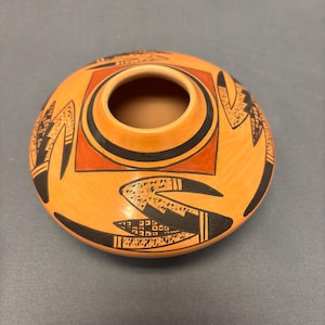 May include: A hand-painted ceramic bowl with a wide, open top. The bowl is orange with black and red geometric designs. The rim and interior are a darker brown. The design includes a square and curved lines.