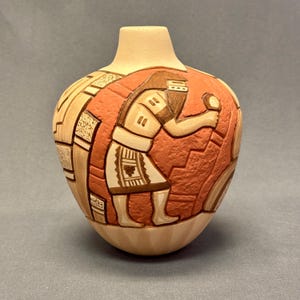 May include: A ceramic vase with a beige neck and base, and a rounded body. The body features a detailed relief design in shades of orange, brown, and beige, depicting a figure holding an object. The design includes geometric patterns and a stepped background.
