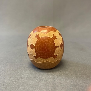May include: A small, round, brown vase with a carved geometric pattern. The vase features a series of star-like shapes and a textured, cracked pattern. The top opening is a darker shade of brown.