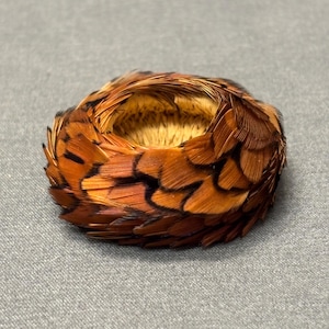 May include: A circular decorative item made of brown and black feathers. The feathers are arranged in a layered pattern, creating a textured appearance. The center of the item is a lighter tan color. The item is likely a decorative accent or a craft supply.