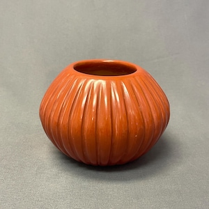 May include: A round, terracotta-colored ceramic vase with a wide, open mouth and a smooth, glossy finish. The exterior surface features a series of vertical, evenly spaced ridges that extend from the base to the rim, creating a textured appearance.