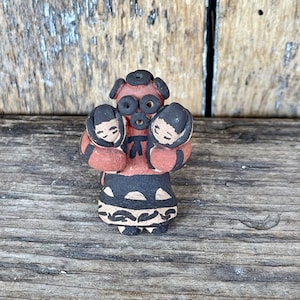 May include: Hand-painted ceramic figurine of a woman carrying two children. The woman is wearing a red dress with black and white geometric patterns. The children are wearing brown and black clothing. The figurine is made of clay and has a rustic, handmade look.