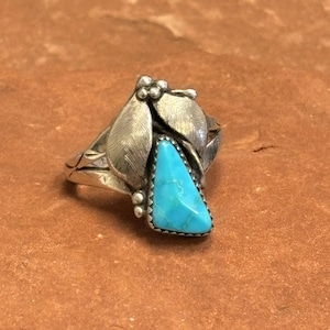 May include: A silver ring featuring a triangular turquoise stone. The ring has a leaf design on the sides and top, with small beaded accents. The turquoise stone is a vibrant blue color, set in a silver bezel.