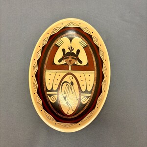 May include: Oval-shaped ceramic bowl with a cream-colored exterior and a central design. The design features a brown and black oval with a cream-colored figure and geometric patterns. The bowl is likely handmade.