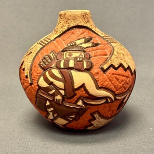 May include: A handcrafted, round ceramic vase with a textured surface. The vase features a detailed relief of a Native American figure in shades of brown, beige, and red. The top and rim of the vase are a lighter beige color.