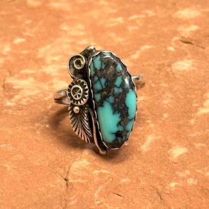 May include: A silver ring featuring a large, oval turquoise stone with black matrix. The ring has decorative silver accents, including a spiral, a circular design, and a feather. The turquoise stone is a vibrant teal color.