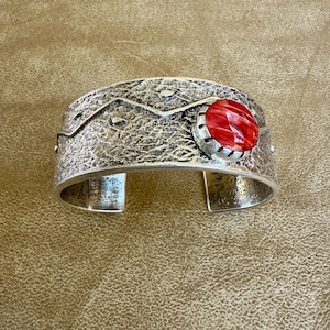 May include: Silver cuff bracelet with a textured surface and a vibrant red gemstone. The bracelet features a mountain-like design along the top edge and is open at the bottom. The gemstone is round and set in a silver bezel.