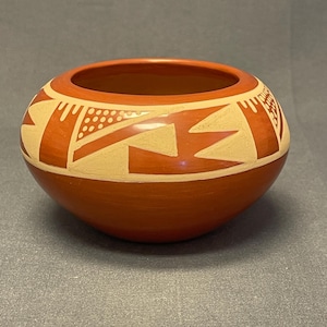 May include: A round, terracotta-colored pottery bowl with a wide opening. The bowl features a cream-colored geometric design around the rim, including zigzags, dots, and stylized shapes. The interior of the bowl is a darker shade of terracotta.