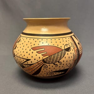 May include: A hand-painted ceramic pot with a beige base and a black rim. The pot features a stylized bird design in shades of brown and black, with a cross-hatch pattern on the bird's body. The background is decorated with small black crosses.